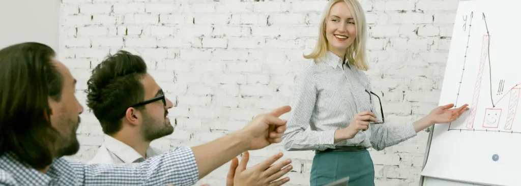 Saleswoman presenting a rising sales chart on a flipchart to two engaged colleagues, illustrating successful results from a Challenger selling strategy in a downturn.